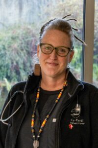 A smiling woman with light skin and glasses stands indoors. She has dreadlocks tied up, wears large wooden earrings, a black jacket with a stethoscope, and a colorful beaded necklace. The background shows greenery through a window.