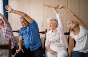 Four older adults sit on chairs in a row, smiling and stretching their arms overhead to one side in a gentle group exercise. They appear relaxed and engaged, wearing casual clothes, with a bright indoor background.