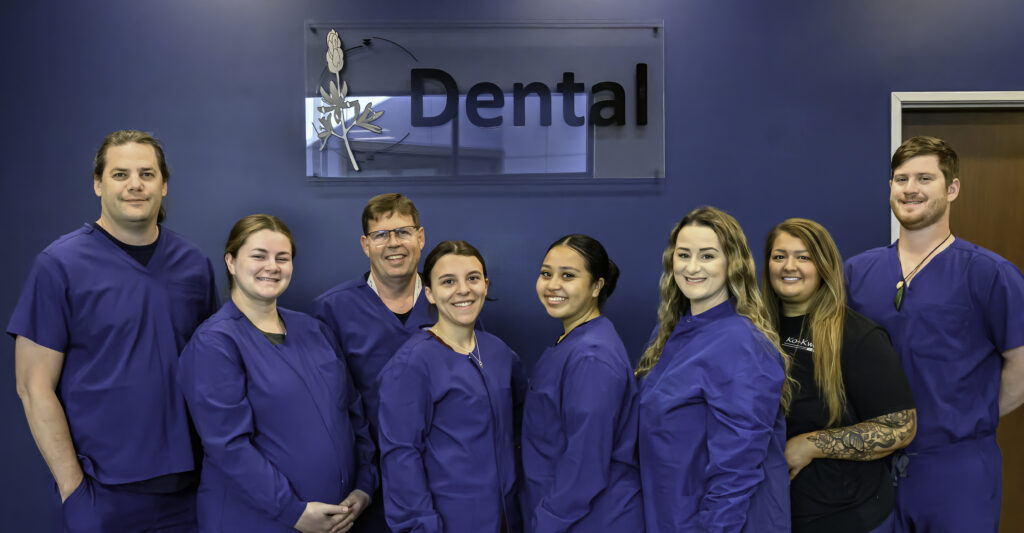 Eight people wearing blue dental scrubs stand smiling in front of a blue wall with a sign that reads “Dental.” One person at the far right wears a black shirt with tattoos visible on their arm. The group appears diverse in age and gender, all facing the camera.