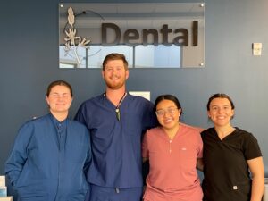 Four dental professionals pose smiling in front of a blue wall with a sign that reads Dental. Three women—one in blue, one in pink, one in black scrubs—stand beside a man in blue scrubs, all looking at the camera in a clinic or office setting.