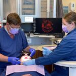 Two dental professionals in blue scrubs, masks, and gloves attend to a patient at KWC-Coos Bay. The patient’s mouth is open for examination, with dental tools nearby and a computer screen displaying the Coquille Indian Tribe logo.