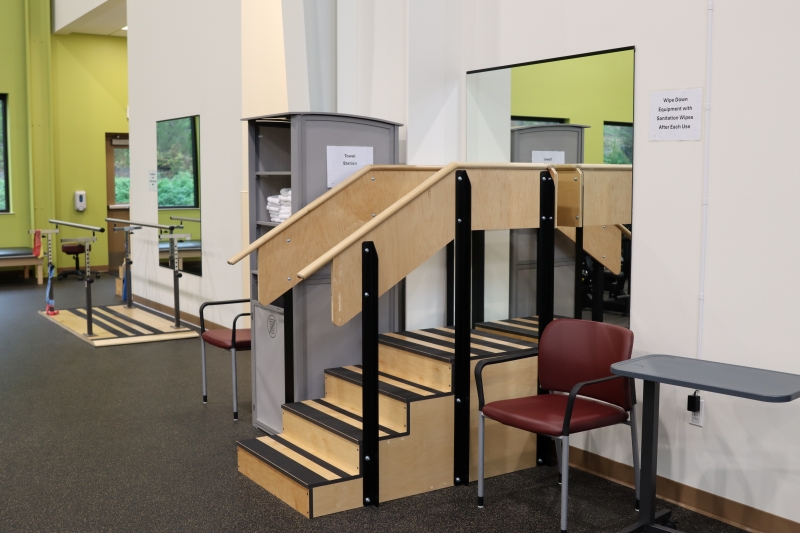 A physical therapy area designed for rehabilitation, featuring parallel bars, two sets of wooden training stairs with handrails, mirrors, a maroon chair with a tray, and a storage cabinet stocked with neatly folded towels.