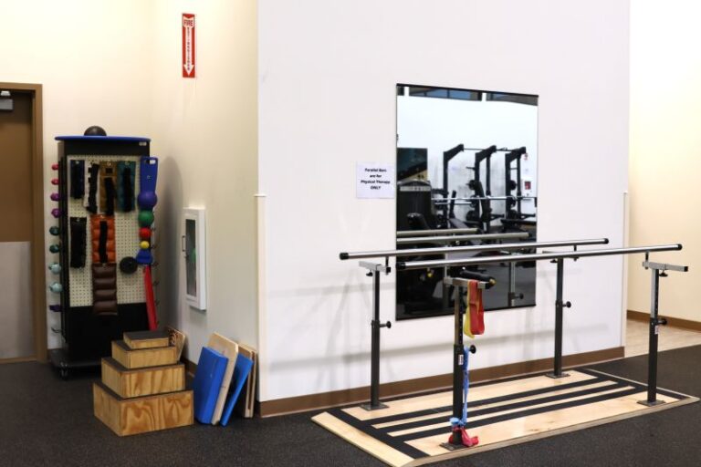 A physical therapy gym area with wooden steps, balance bars, and therapy bands. A mirrored wall reflects equipment in the background. To the left, a rack holds exercise balls, weights, and resistance bands for rehabilitation exercises. Blue foam pads rest by the steps.