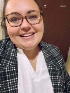 A person with wavy brown hair, wearing tortoiseshell glasses, a green checkered blazer, and a white shirt is smiling at the camera indoors. The background shows a brown door with a white door number.