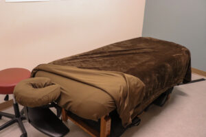 A massage table covered with a brown blanket and tan sheet is set up in a room with light-colored walls and flooring. A headrest is attached, and a round red stool and black chair are positioned nearby. The room appears calm and organized.
