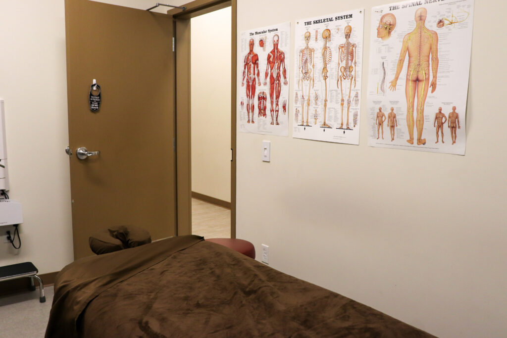 A massage therapy room at Shishda Haws features a brown, covered table in the foreground. The door is open, with anatomical charts of muscles, the skeleton, and the nervous system displayed above a light switch.