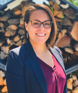 A woman with short brown hair and glasses smiles outdoors. She wears a navy blazer over a burgundy blouse and earrings. Stacked firewood is visible in the background, and sunlight highlights her face.