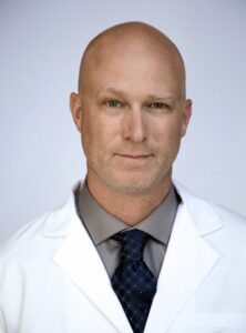 A bald man wearing a white lab coat, grey shirt, and dark blue patterned tie, stands facing the camera against a plain light background. He has light skin, blue eyes, and a neutral expression. This image appears to be a professional or medical portrait.