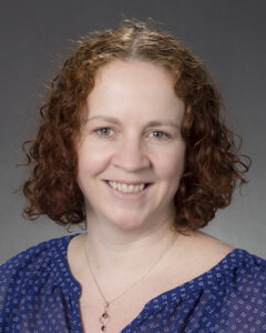 A woman with fair skin and shoulder-length, curly auburn hair smiles at the camera. She is wearing a blue blouse with a subtle white pattern and a delicate necklace. The background is a neutral gray gradient.