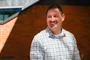A man with short brown hair and a mustache, wearing a white and gray plaid shirt, smiles while standing outside against a background of red brick walls and diagonal shadows—a true community connector, warmed by bright sunlight.