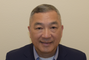A middle-aged man with short gray hair smiles at the camera. He is wearing a dark blazer over a light blue collared shirt. The background is a plain, light beige wall.