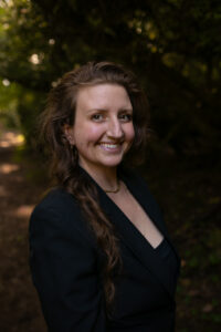 A woman with long, wavy brown hair smiles at the camera. She wears a black blazer, a black top, and gold jewelry. The background shows a wooded outdoor setting with sunlight filtering through the trees and a dirt path behind her.