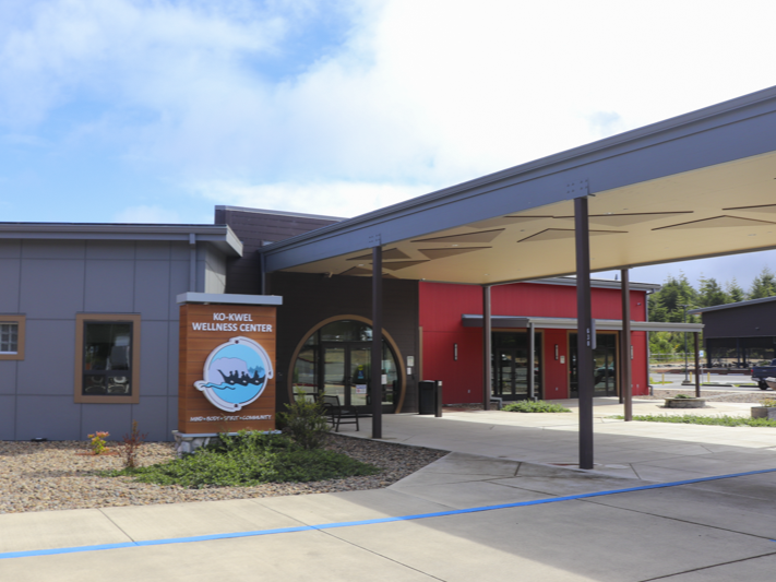 Modern building with gray and red walls, large windows, and an overhang entrance. A sign in front reads Ko-Kwel Wellness Center with a blue wave and canoe logo. Sidewalks and landscaped areas surround the entrance.