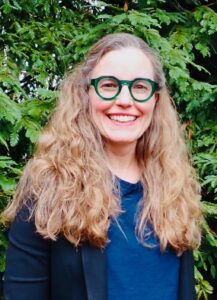 A person with long wavy blonde hair, wearing green glasses, a blue top, and a dark blazer, smiles in front of lush green foliage.