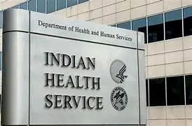 A metal sign reads Indian Health Service with two official logos and Department of Health and Human Services above. The background features windows and a concrete office building facade, highlighting the IHS’s commitment to Nutrition Initiatives and Tribal Food programs within Native communities.