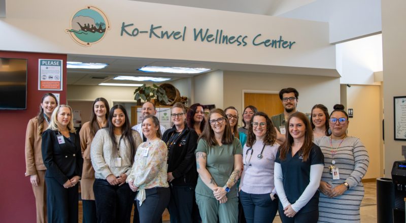 The 16-person team of KWC Eugene stands together in a lobby under a sign that reads “Ko-Kwel Wellness Center.” Some wear professional attire, others wear scrubs or office clothes. The setting looks welcoming, with office decor and natural light.