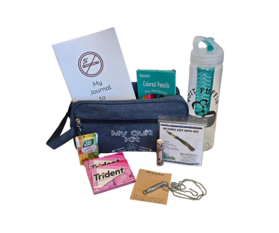A quit smoking kit containing a denim zip pouch labeled My Quit Kit, a water bottle, a journal, colored pencils, a Breathe necklace on a chain, a Trident gum pack, a stress relief mints tin, and a beaded key ring kit, all arranged neatly.